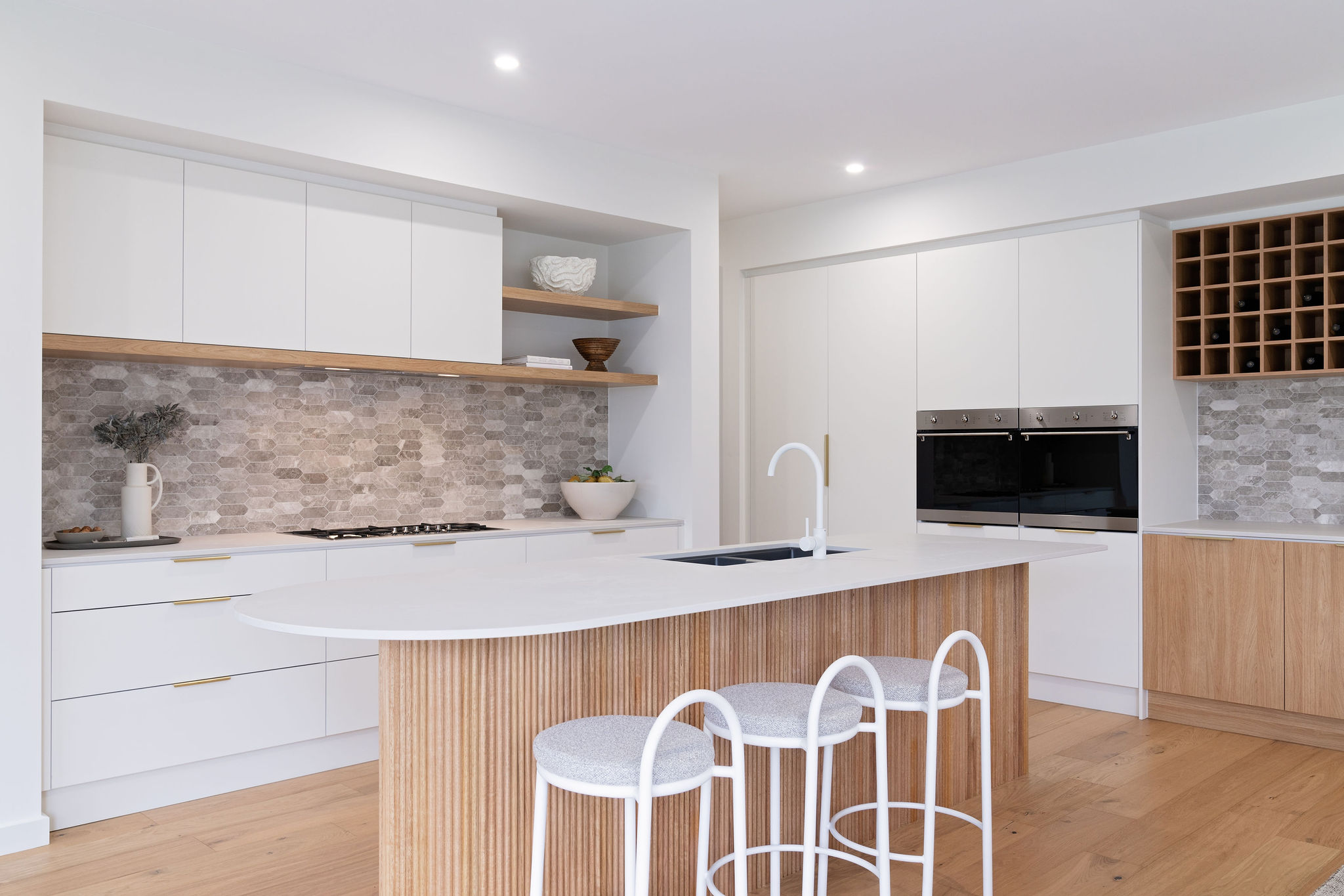 A modern display kitchen with white cabinetry, a large island bench, pendant lighting, and stainless steel appliances, showcasing the design of a display home.